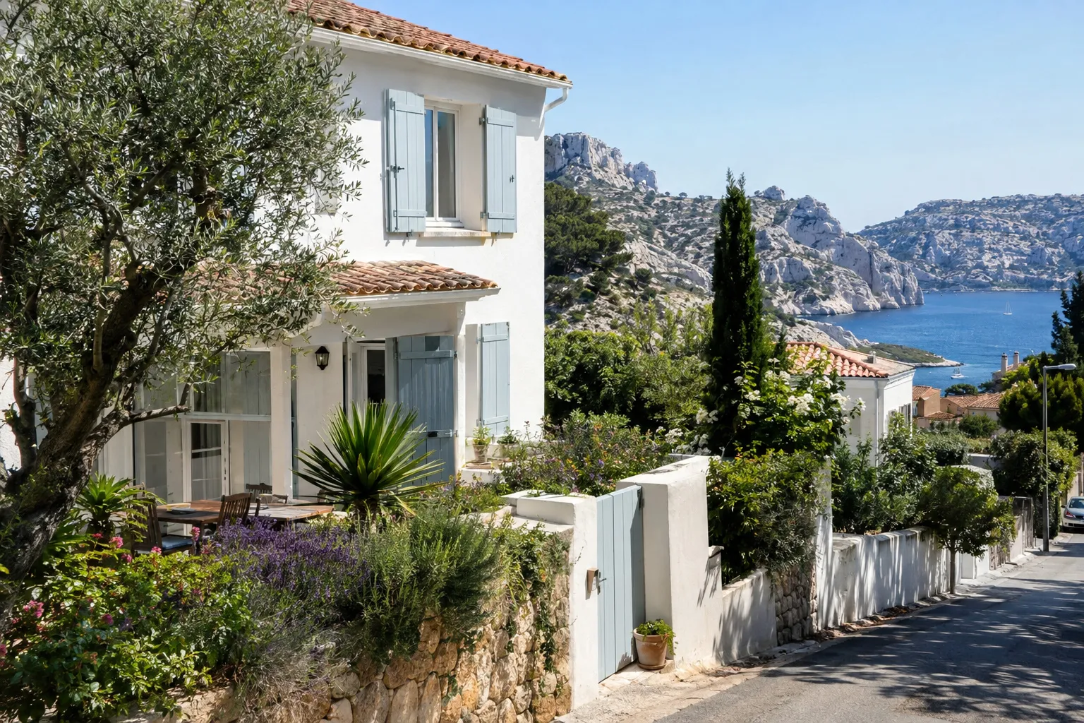 Maison blanche à volets bleu-gris et toit de tuiles dans une rue en pente du sud de Marseille, avec un olivier, de la lavande et une calanque au fond.